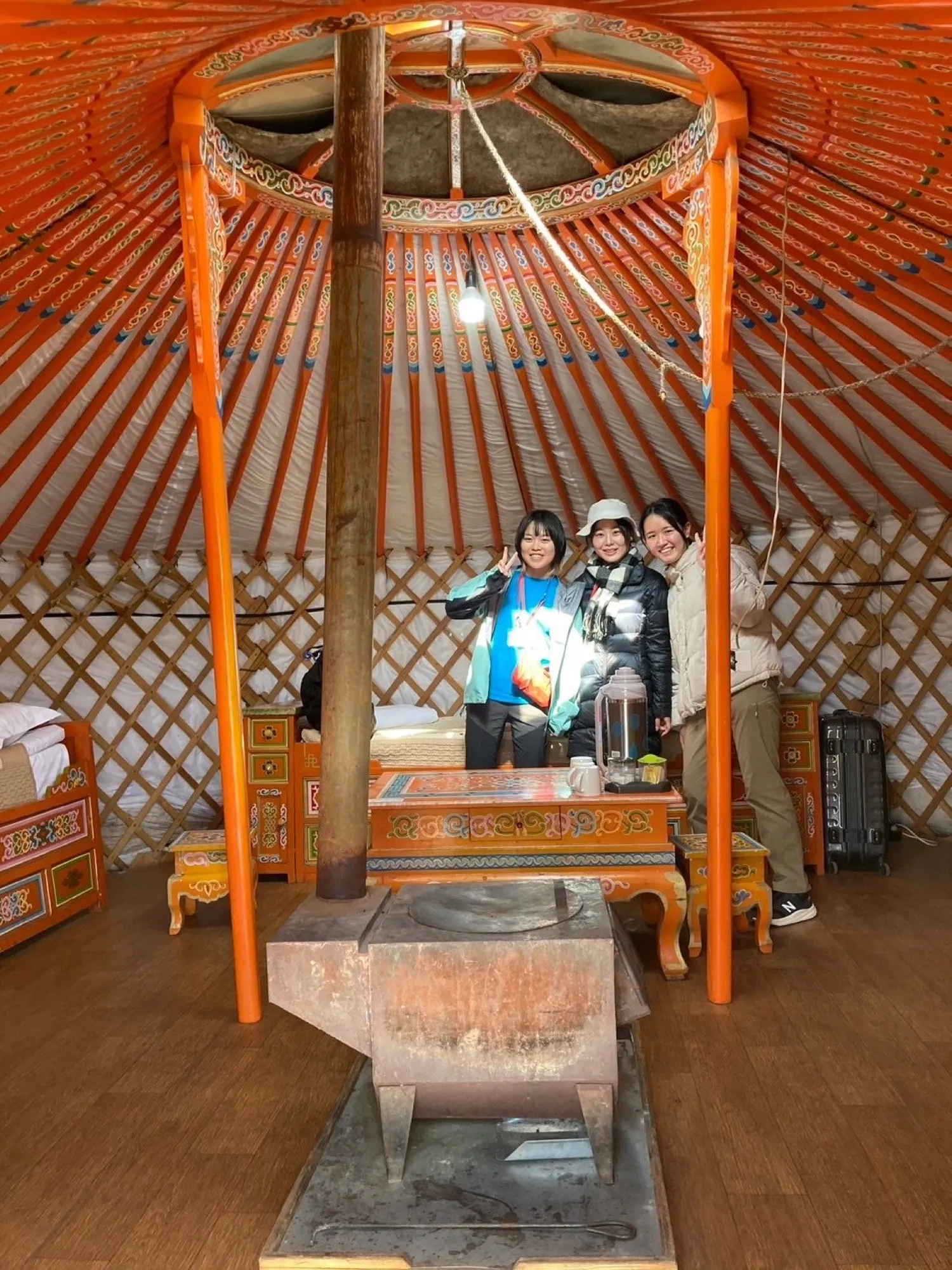 Inside a ger: an orange wooden roof frame, a stove in the centre and three people standing at the back.