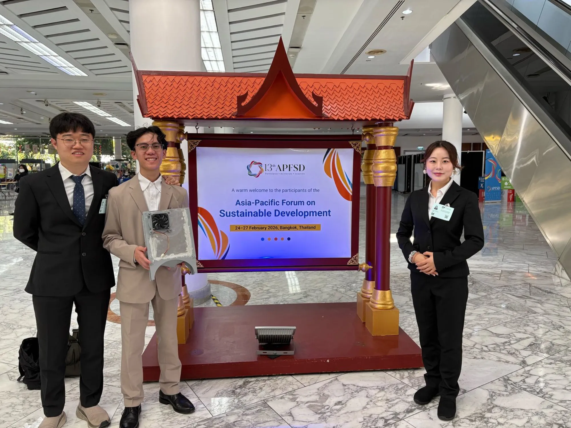 Three HydroBeetle team members at the Asia-Pacific Forum on Sustainable Development in Bangkok, one holding a collecting panel, beside the forum’s welcome sign.