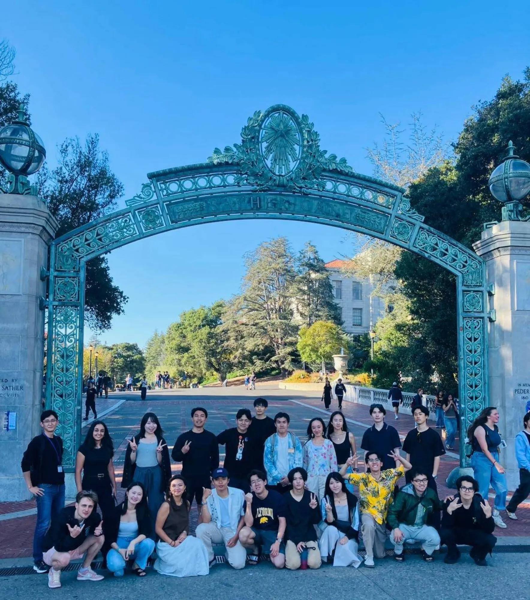 A large group of programme participants posing in front of an ornate green gateway on a sunny campus.