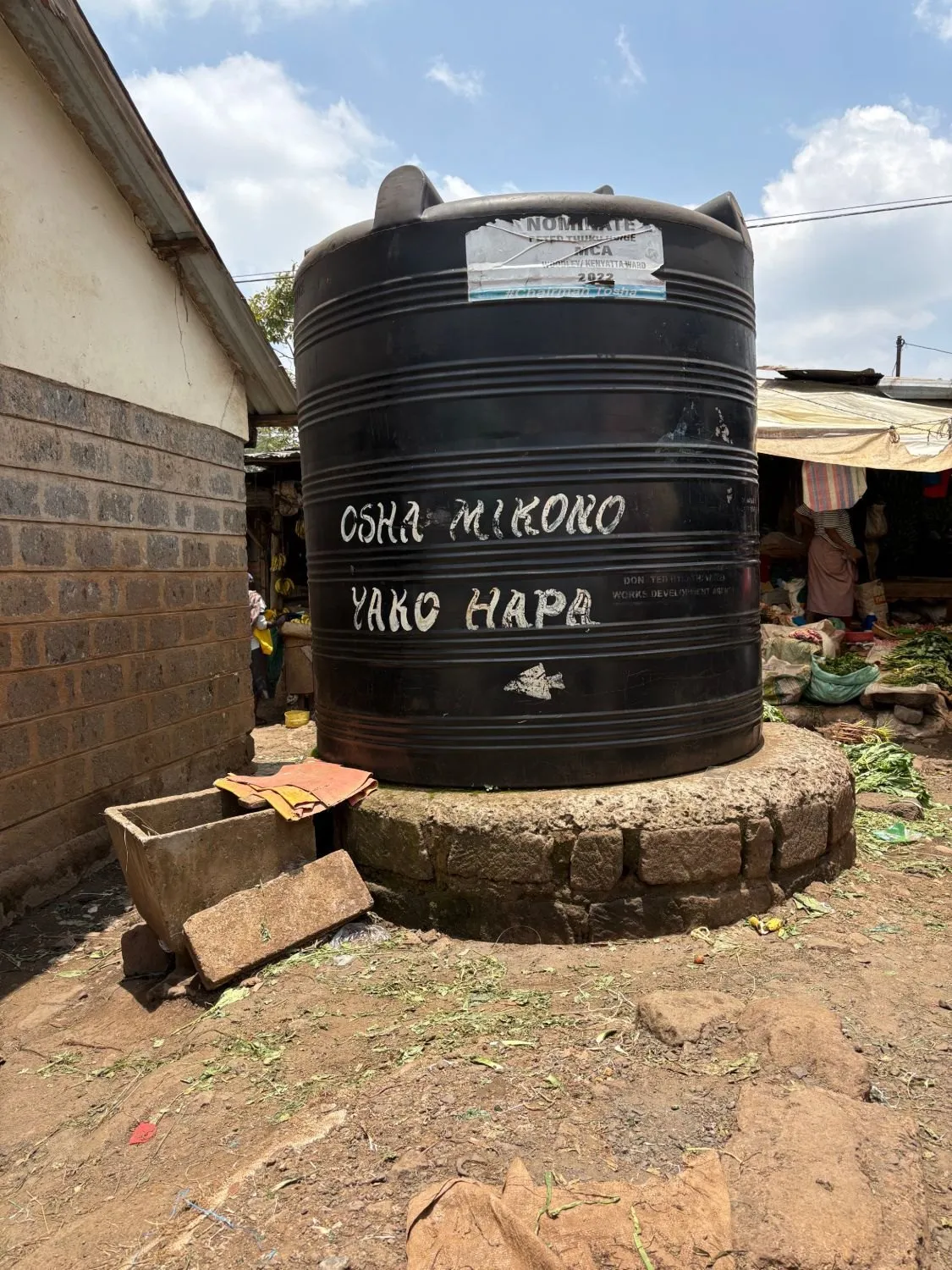 A large black water tank at a market, painted with the words “Osha mikono yako hapa”.